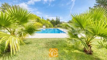 Gemeenschappelijk buitenzwembad bij Villa Lloret, omringd door groene palmbomen, een grasgazon en bomen onder een blauwe hemel.