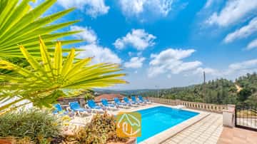 Privézwembad met blauwe ligbedden en een tak van een palmprom op de voorgrond bij Villa Santa Maria onder een blauwe lucht met her en der wat wolken.