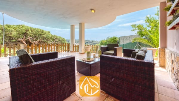 Overdekt terras bij Villa Valentina met bruine rieten fauteuils, een salontafel met glazen blad en tegelvloeren die uitkijken op groene bomen en een blauwe lucht.