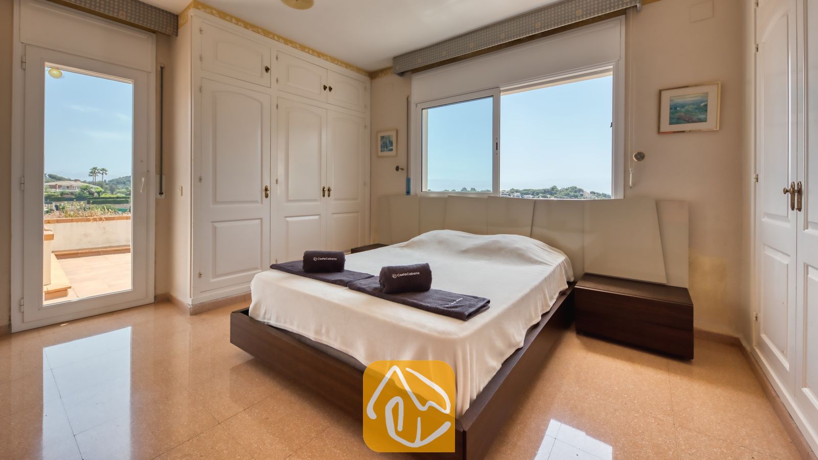 Slaapkamer met tweepersoonsbed, donkerhouten ombouw, witte kledingkasten, raam met uitzicht op bomen en de lucht, en een balkondeur.