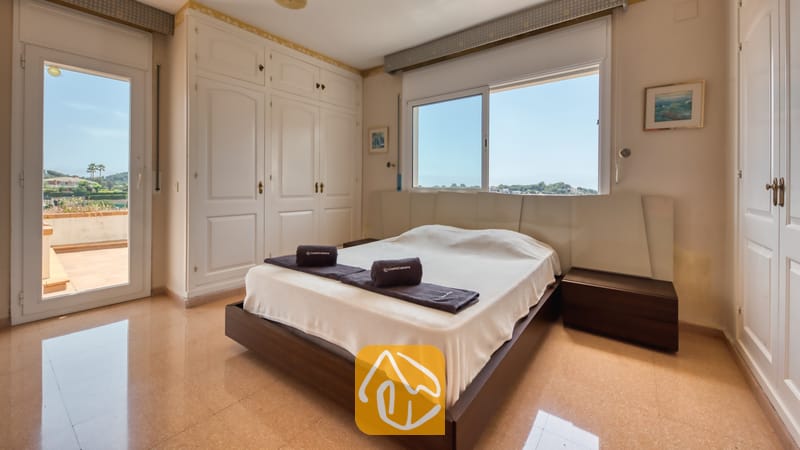 Slaapkamer met tweepersoonsbed, donkerhouten ombouw, witte kledingkasten, raam met uitzicht op bomen en de lucht, en een balkondeur.