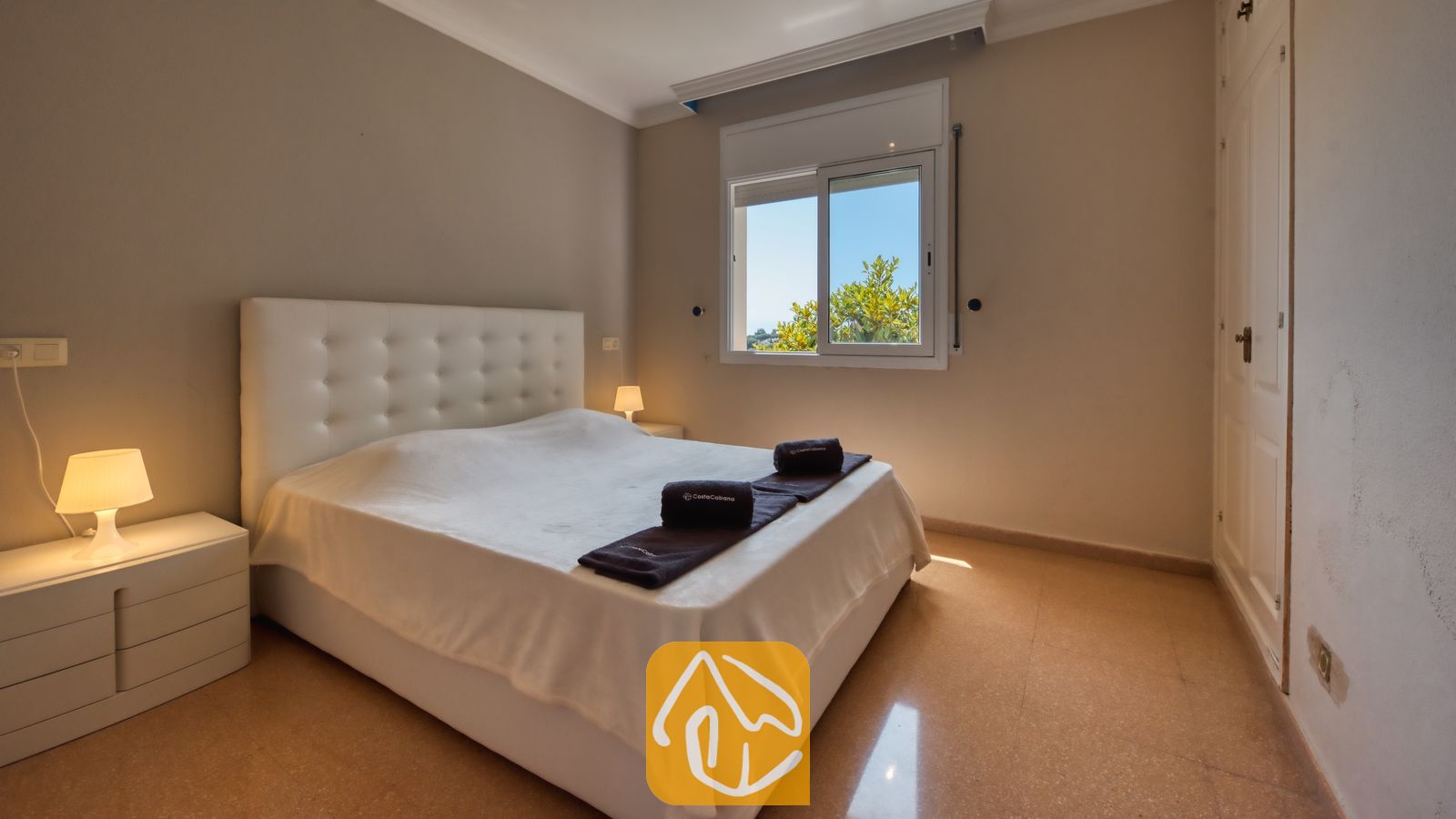 Slaapkamer met een bed met een wit gecapitonneerd hoofdbord, twee nachtkastjes met kleine lampen en een raam dat uitzicht biedt op groen loof tegen een blauwe lucht.