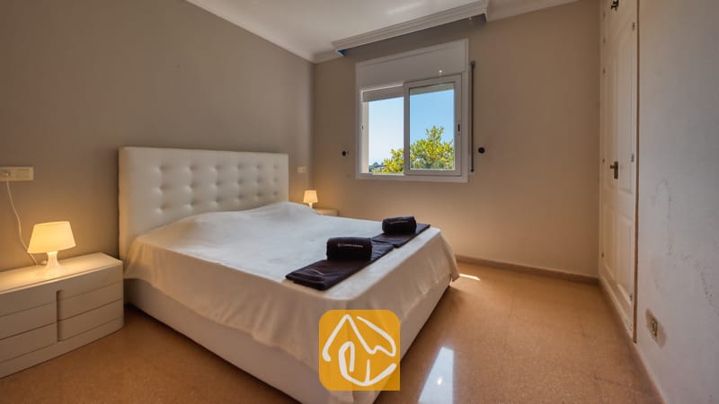 Slaapkamer met een bed met een wit gecapitonneerd hoofdbord, twee nachtkastjes met kleine lampen en een raam dat uitzicht biedt op groen loof tegen een blauwe lucht.