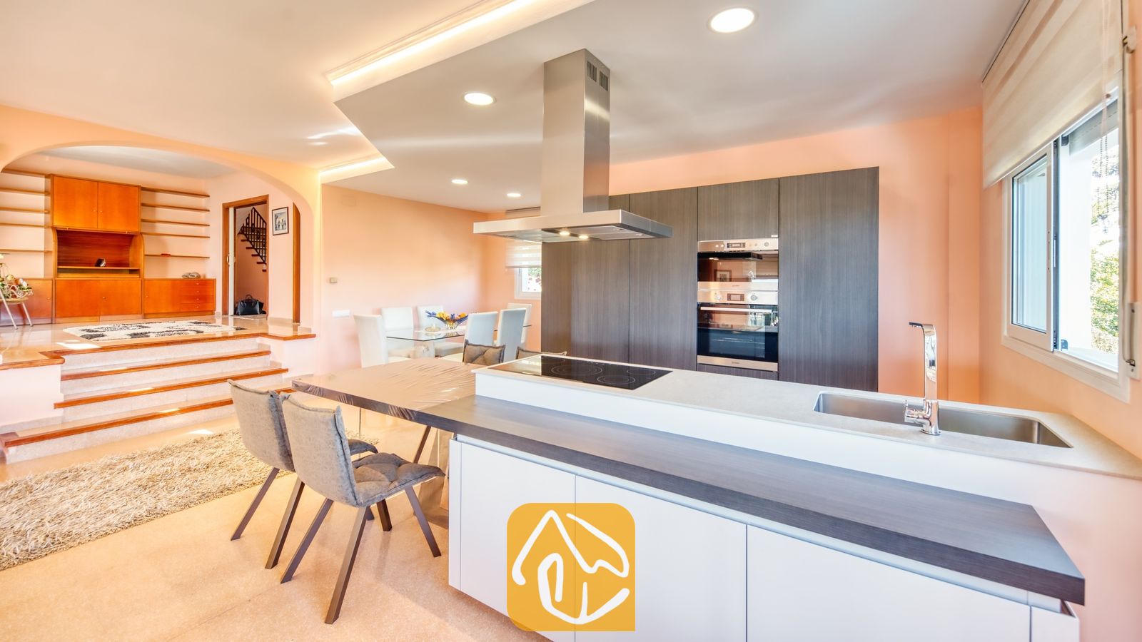 Moderne keuken met donkere houten kasten, een roestvrijstalen afzuigkap en een aangrenzende eethoek met een trap naar een verhoogde woonkamer.