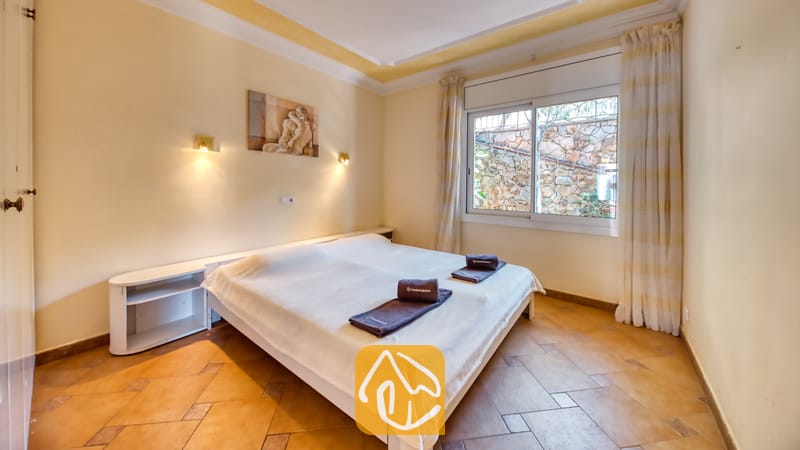 Slaapkamer met een tweepersoonsbed opgemaakt met witte lakens, donkere opgevouwen handdoeken, tegelvloeren en een raam met gordijnen.