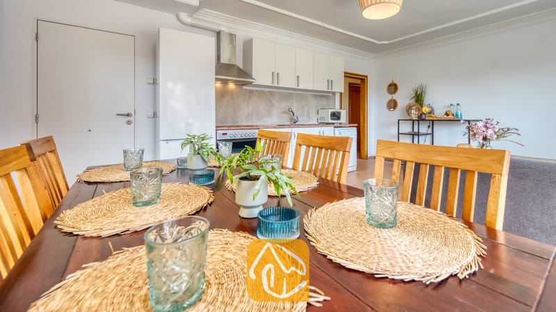 Houten eettafelset met geweven placemats en glaswerk, met een open keuken op de achtergrond.