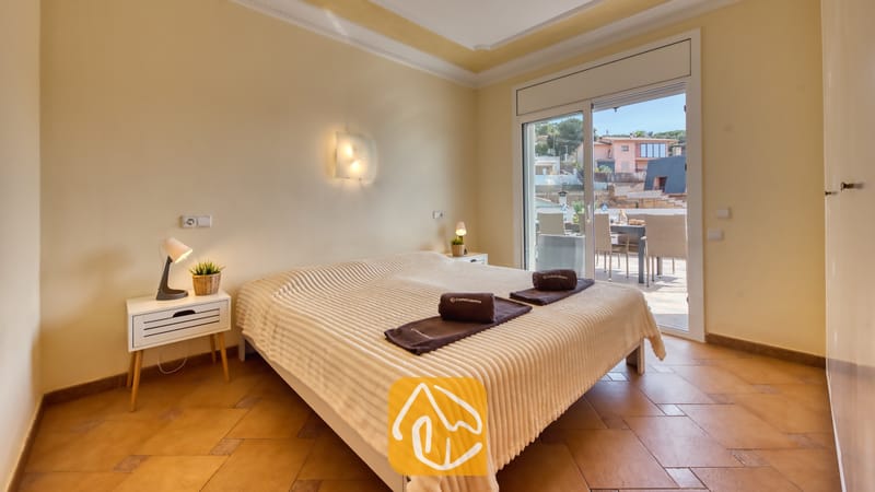 Slaapkamer in Villa Donna met een tweepersoonsbed, een witte geweven sprei, opgerolde donkere handdoeken en glazen schuifdeuren die opengaan naar een buitenterras