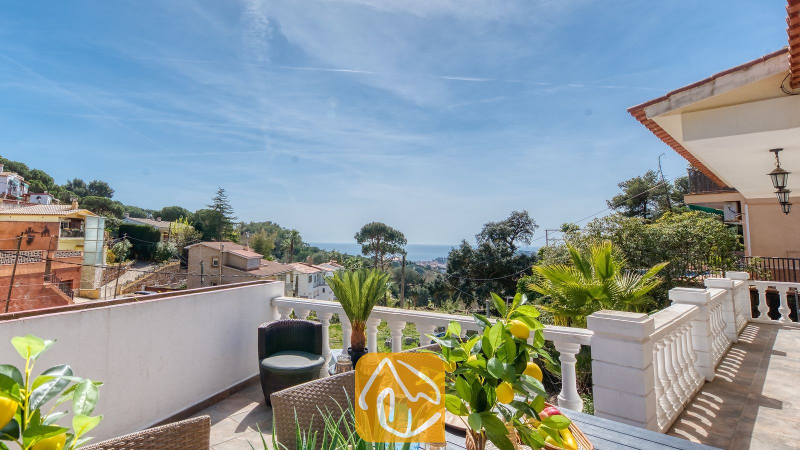 Balkonterras van Villa Donna met uitzicht over groene heuvels en de blauwe zee, voorzien van een eettafel met een in een pot geplante citroenboom en geweven stoelen onder een heldere hemel.
