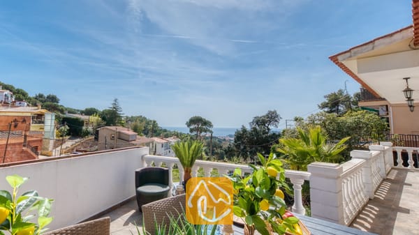 Balkonterras van Villa Donna met uitzicht over groene heuvels en de blauwe zee, voorzien van een eettafel met een in een pot geplante citroenboom en geweven stoelen onder een heldere hemel.