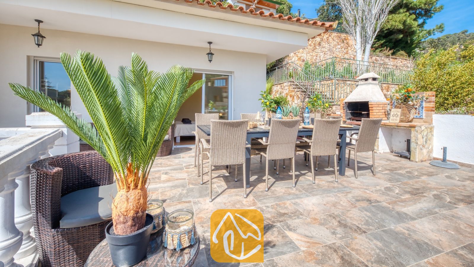 Zonnig terras bij Villa Donna, voorzien van een eettafel voor buiten met rieten stoelen, een ingebouwde stenen barbecue en betegelde vloeren.