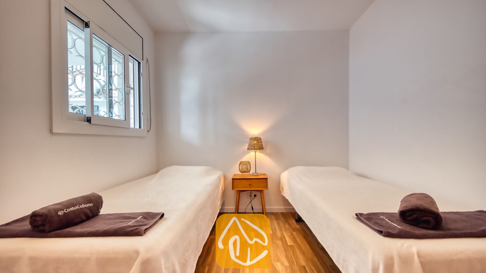 Slaapkamer met twee eenpersoonsbedden, voorzien van wit beddengoed, donkerbruine opgerolde handdoeken, een houten nachtkastje en een klein tafellampje.