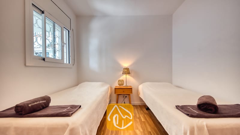 Slaapkamer met twee eenpersoonsbedden, voorzien van wit beddengoed, donkerbruine opgerolde handdoeken, een houten nachtkastje en een klein tafellampje.