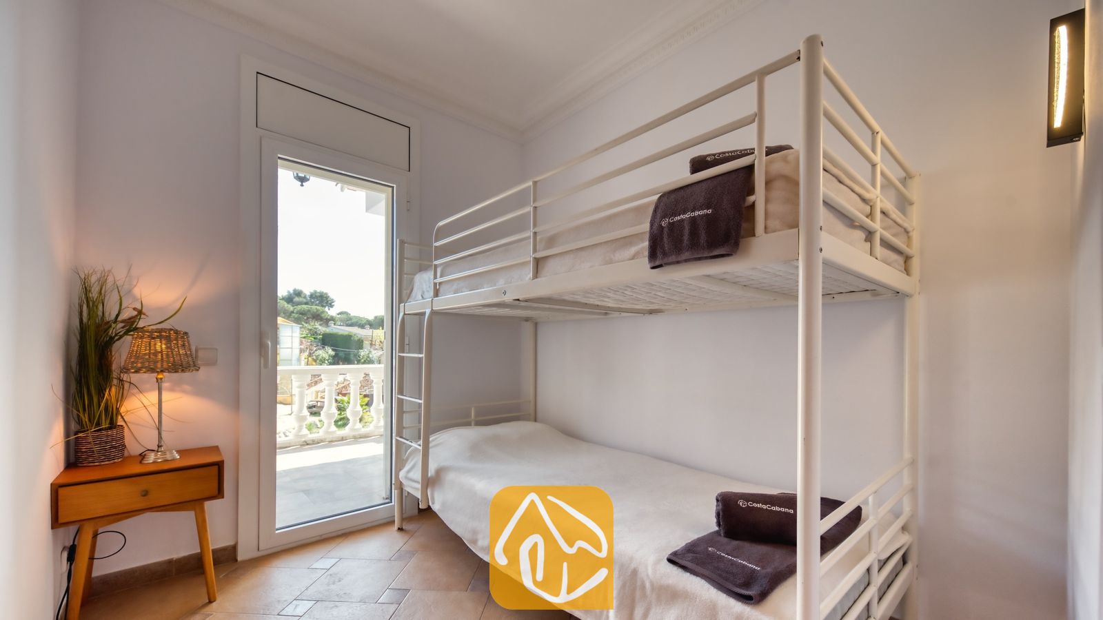 Slaapkamer in Villa Donna met een wit metalen stapelbed voorzien van opgevouwen donkere handdoeken, een houten nachtkastje met een gevlochten lamp en een glazen deur die uitkomt op een balkon.