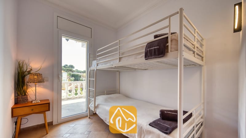 Slaapkamer in Villa Donna met een wit metalen stapelbed voorzien van opgevouwen donkere handdoeken, een houten nachtkastje met een gevlochten lamp en een glazen deur die uitkomt op een balkon.