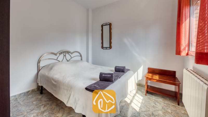 Slaapkamer met een wit bed, grijs gevouwen handdoeken, een metalen hoofdbord, rode gordijnen en een houten bagagerek.