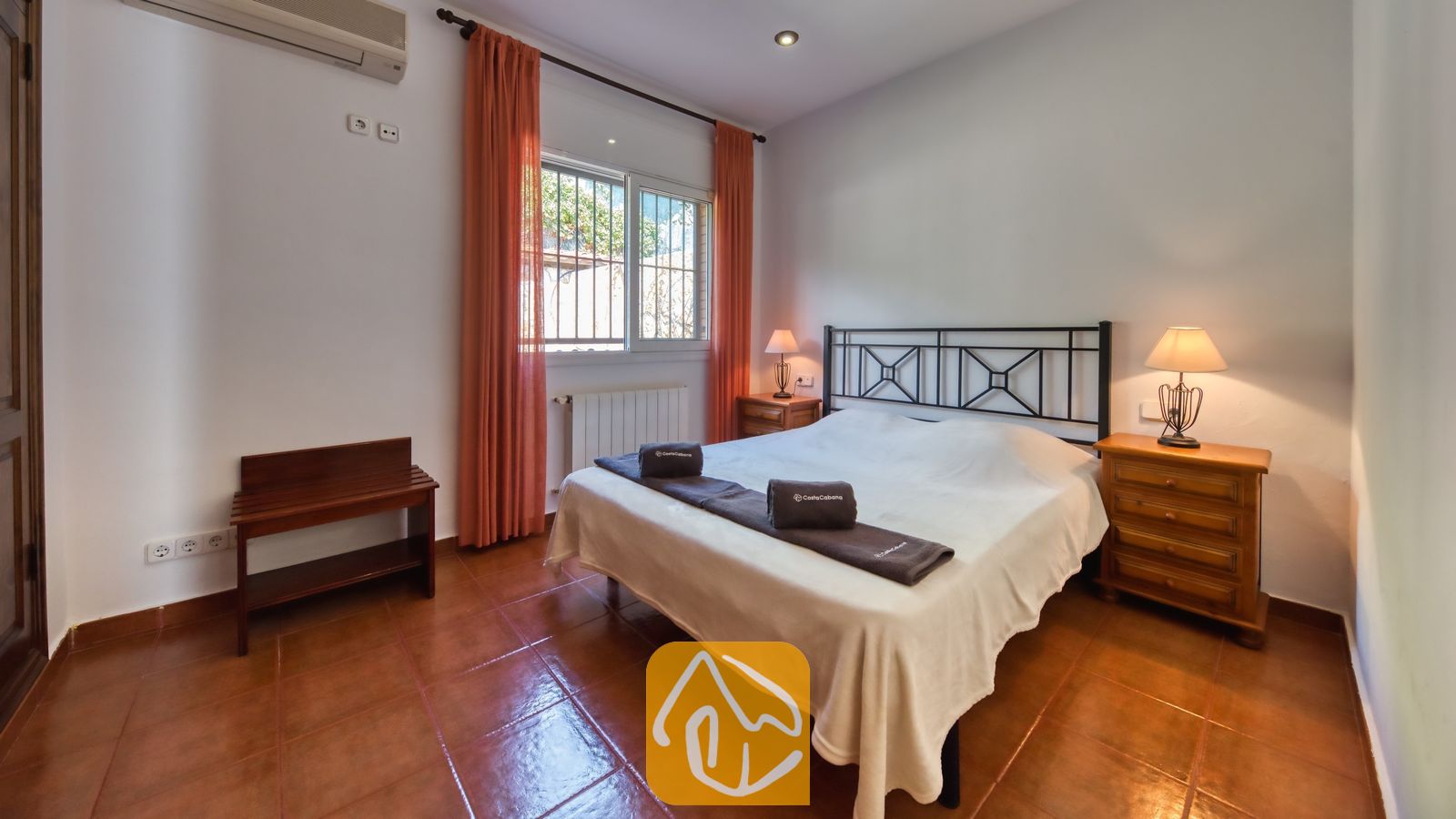 Slaapkamer met een tweepersoonsbed, een donker metalen frame, oranje gordijnen die het raam omlijsten, en twee houten nachtkastjes met lampen.