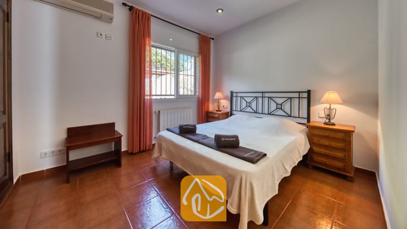 Slaapkamer met een tweepersoonsbed, een donker metalen frame, oranje gordijnen die het raam omlijsten, en twee houten nachtkastjes met lampen.