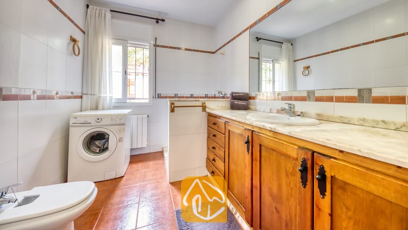 Lichte, betegelde badkamer met een houten badkamermeubel met een marmeren blad, een witte wasmachine en terracotta vloertegels.