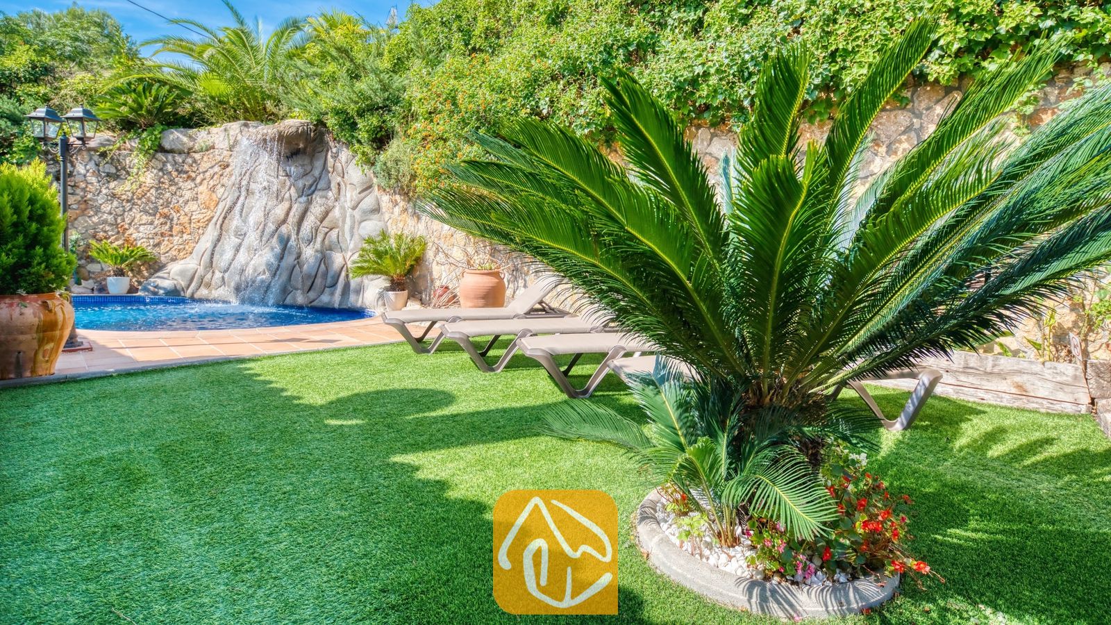 Weelderige groene tuin bij Villa Alba met kunstgras, een opvallende cycadale palm, ligstoelen en een stenen waterpartij met waterval.