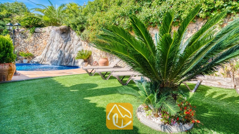 Weelderige groene tuin bij Villa Alba met kunstgras, een opvallende cycadale palm, ligstoelen en een stenen waterpartij met waterval.