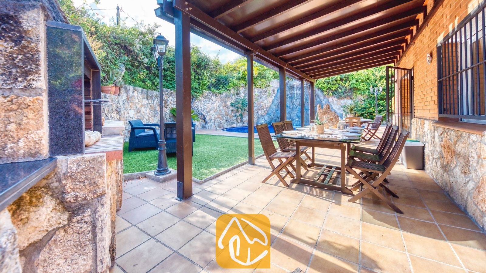 Overdekt buitenterras bij Villa Alba, voorzien van een houten eettafel met stoelen, een tegelvloer en een aangrenzende, ingebouwde barbecuehoek met uitzicht op de tuin en het zwembad.