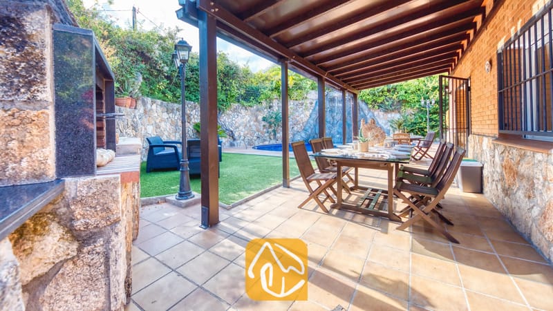 Overdekt buitenterras bij Villa Alba, voorzien van een houten eettafel met stoelen, een tegelvloer en een aangrenzende, ingebouwde barbecuehoek met uitzicht op de tuin en het zwembad.
