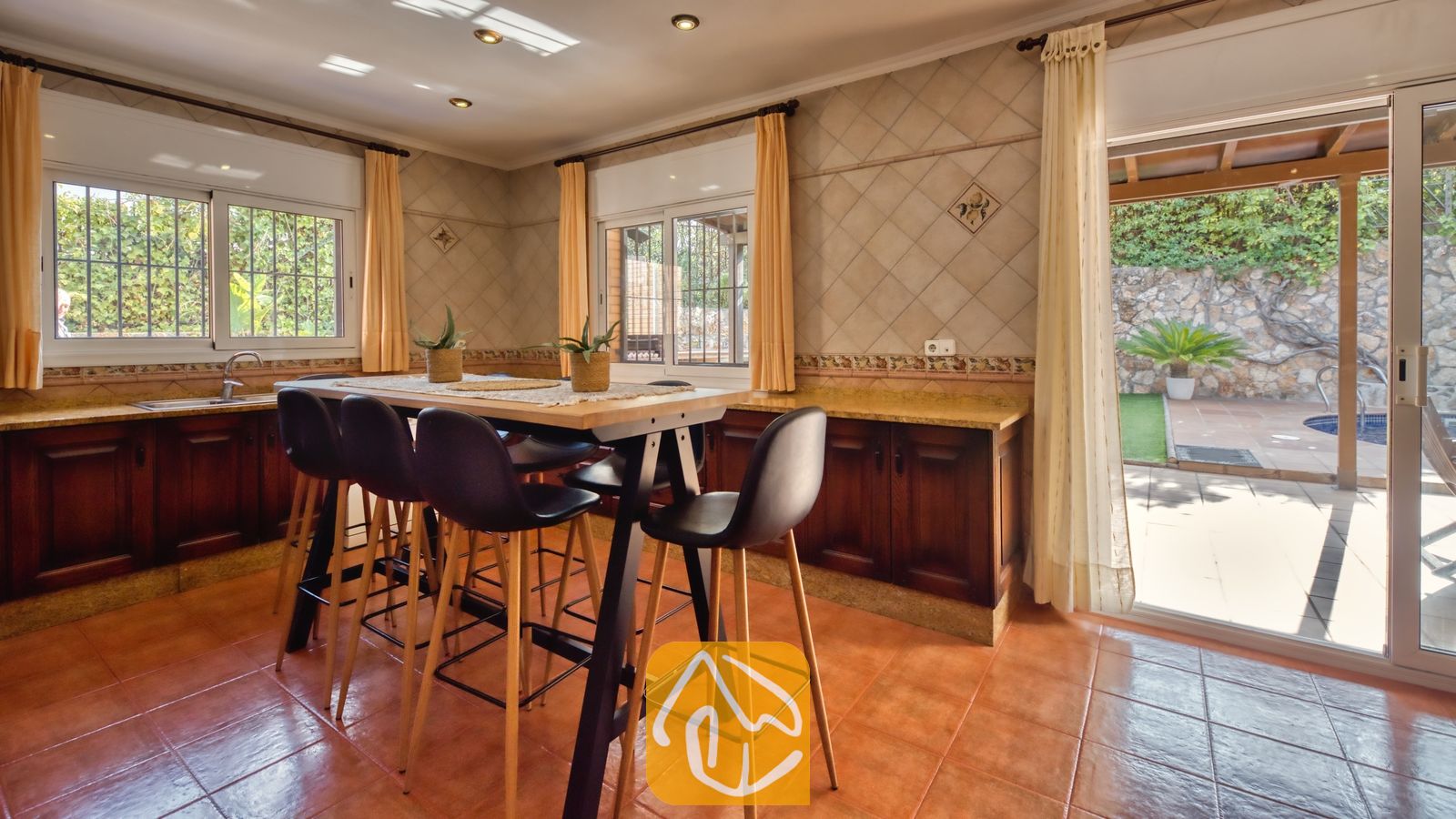 Keuken en eethoek met een hoge houten tafel en zwarte barstoelen, terracotta vloertegels, betegelde muren en een open glazen deur die leidt naar een buitenterras bij Villa Alba.