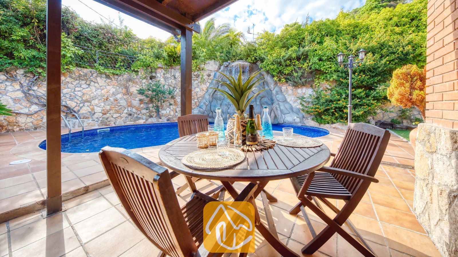 Houten eettafel en stoelen op een stenen terras bij Villa Alba, met een blauw zwembad en een rotswaterval als blikvanger op de achtergrond.