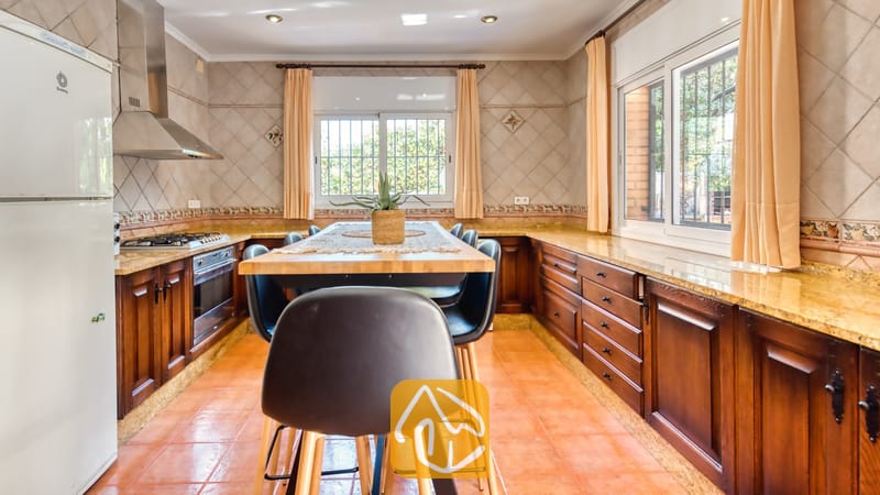Ruime keuken met donkerhouten kasten, granieten aanrechtbladen, een centrale houten eettafel met zwarte stoelen en terracotta vloertegels.
