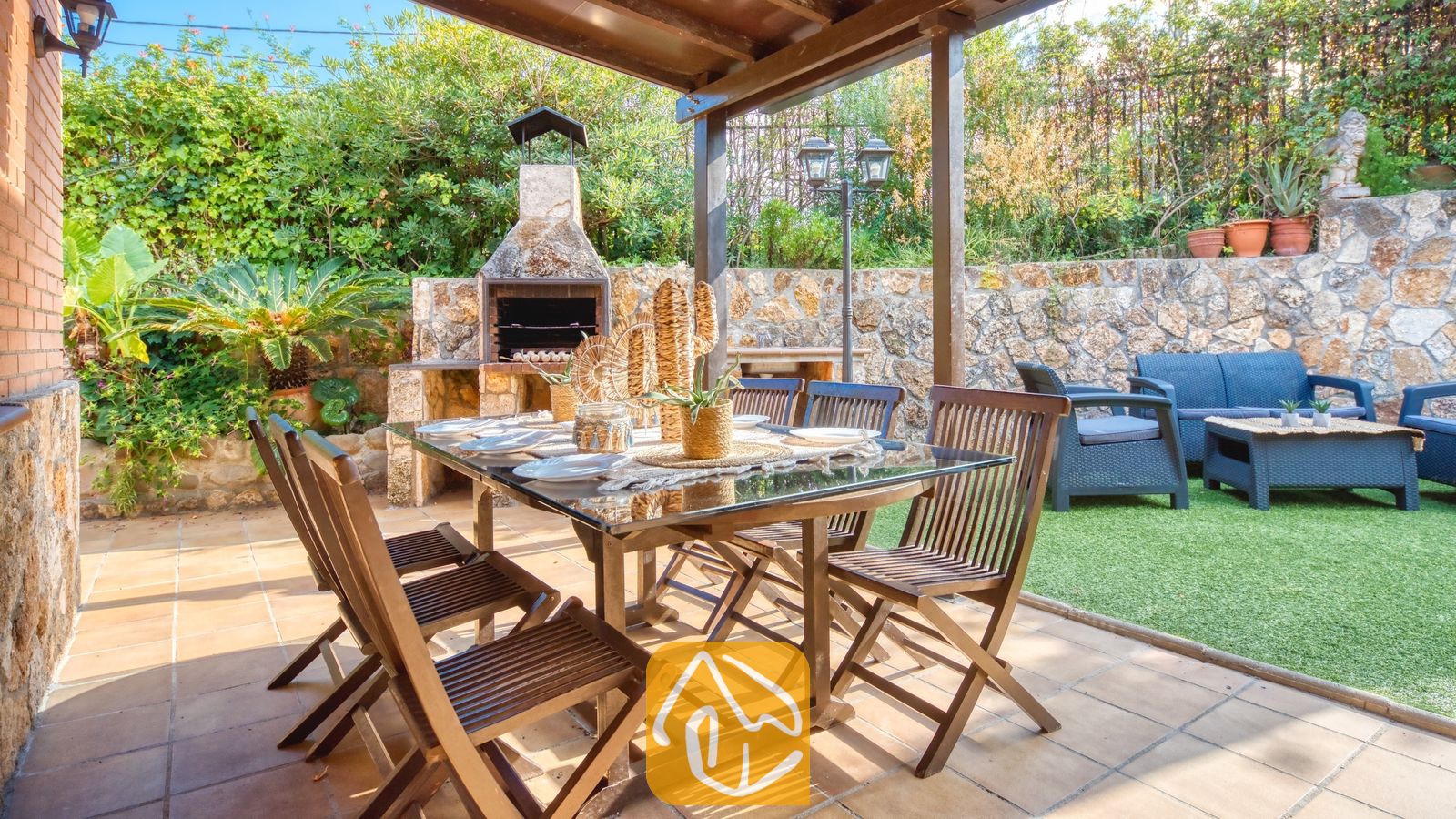 Overdekt buitenbeterterras bij Villa Alba met een houten tafel met glazen blad, klapstoelen en een ingebouwde stenen barbecue op de achtergrond.