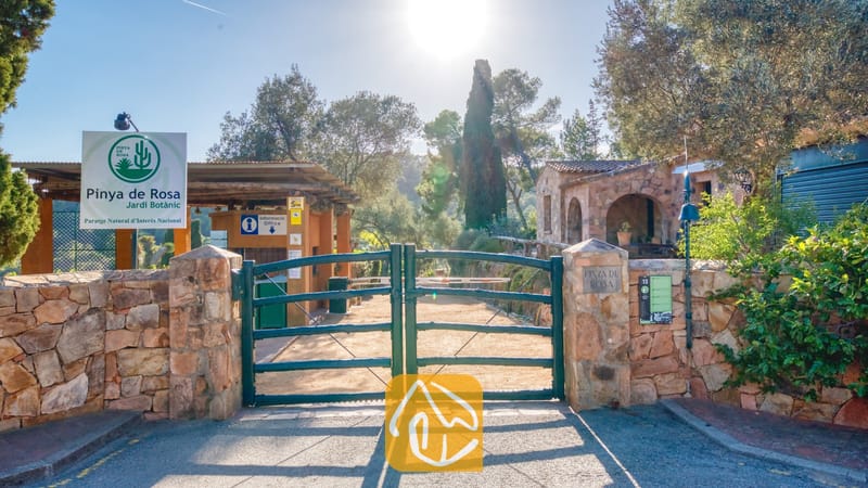 Groene metalen toegangspoort tot de botanische tuin Pinya de Rosa nabij Villa Santa Cristina, gekenmerkt door stenen muren, informatieborden en hoge cipressen in het felle zonlicht.