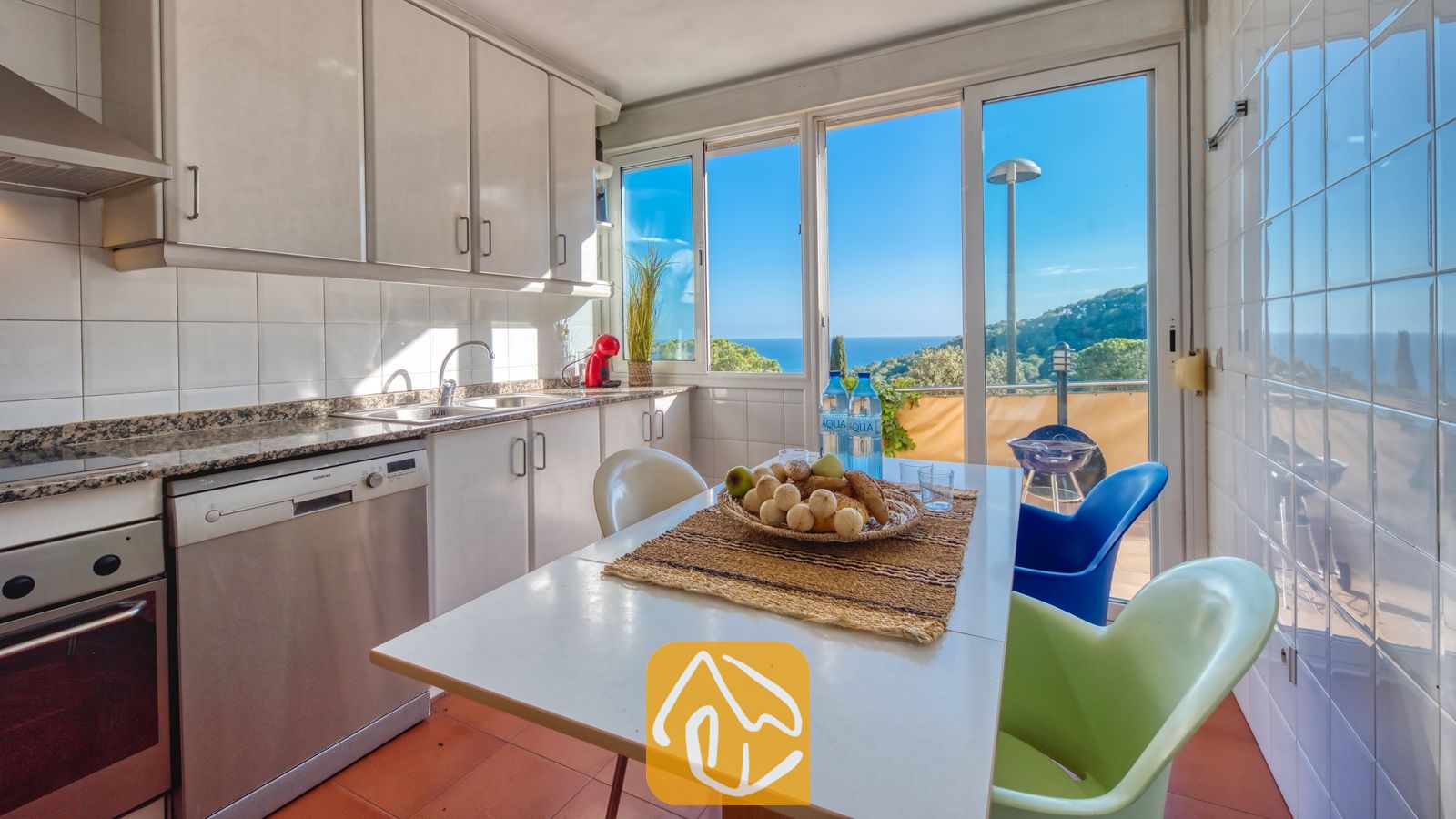 De keuken van Villa Santa Cristina is voorzien van witte kasten, een eettafel met kleurrijke stoelen en een groot raam met uitzicht op de zee en de groene heuvels.