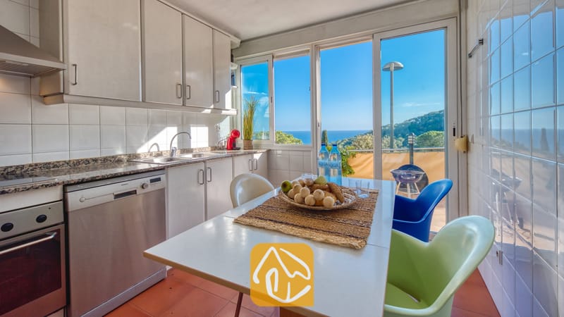 De keuken van Villa Santa Cristina is voorzien van witte kasten, een eettafel met kleurrijke stoelen en een groot raam met uitzicht op de zee en de groene heuvels.
