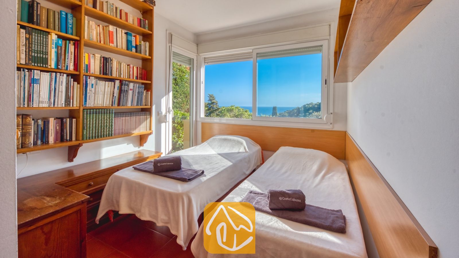 Slaapkamer in Villa Santa Cristina met twee eenpersoonsbedden, wit beddengoed en grijze handdoeken. Naast het raam, dat een prachtig uitzicht op zee biedt, staat een grote houten boekenkast.