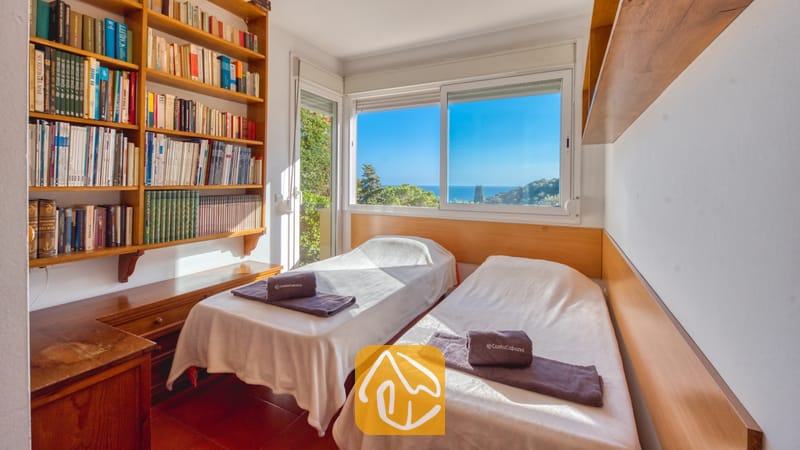 Slaapkamer in Villa Santa Cristina met twee eenpersoonsbedden, wit beddengoed en grijze handdoeken. Naast het raam, dat een prachtig uitzicht op zee biedt, staat een grote houten boekenkast.