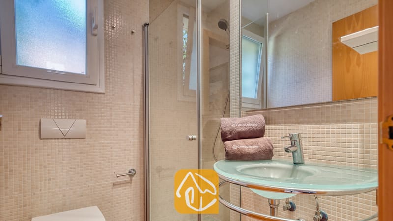 Badkamer met een glazen wastafel op zuil, chromen kraan en een inloopdouche. De wanden zijn betegeld met beige mozaïektegels en een grote spiegel reflecteert het matglazen raam.