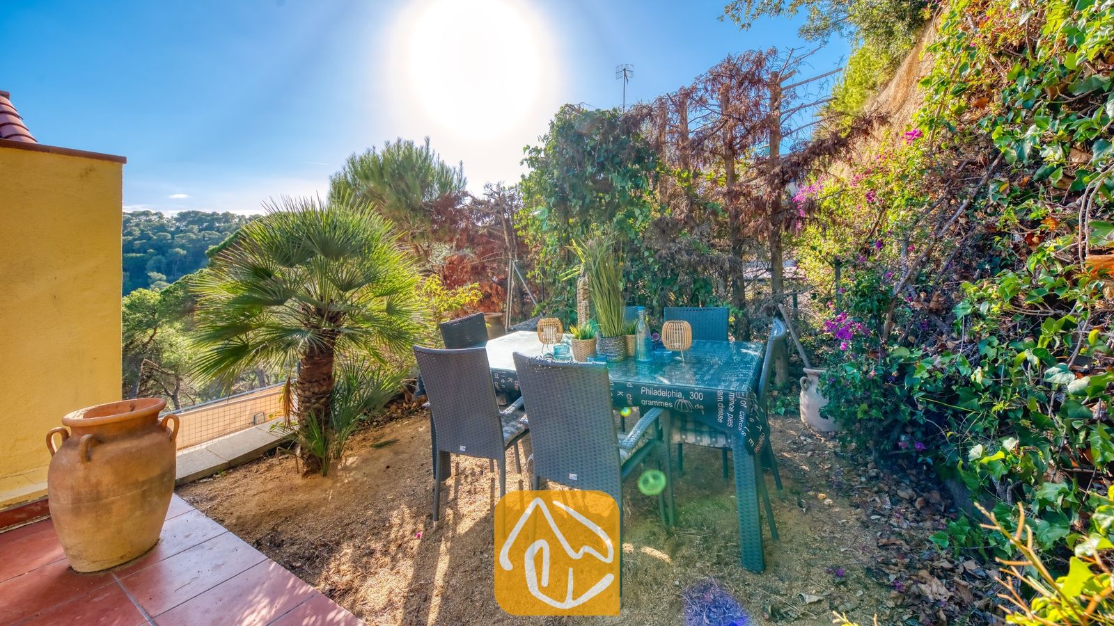 Buitenruimte om te dineren bij Villa Santa Cristina, voorzien van grijze rieten stoelen, een tafel met patroon en een kleine palmboom tegen een achtergrond van een weelderige, groene heuvel.