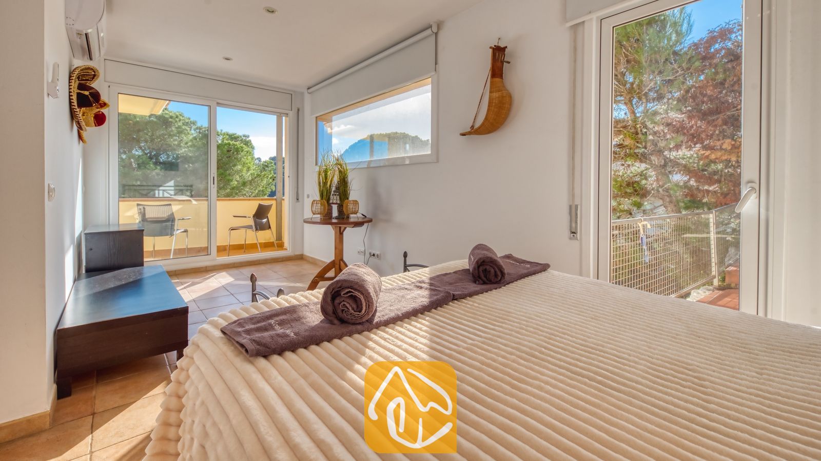 Slaapkamer met een crèmekleurige sprei met textuur en opgerolde paarse handdoeken, voorzien van grote glazen schuifdeuren die uitkomen op een zonnig terras bij Villa Santa Cristina.