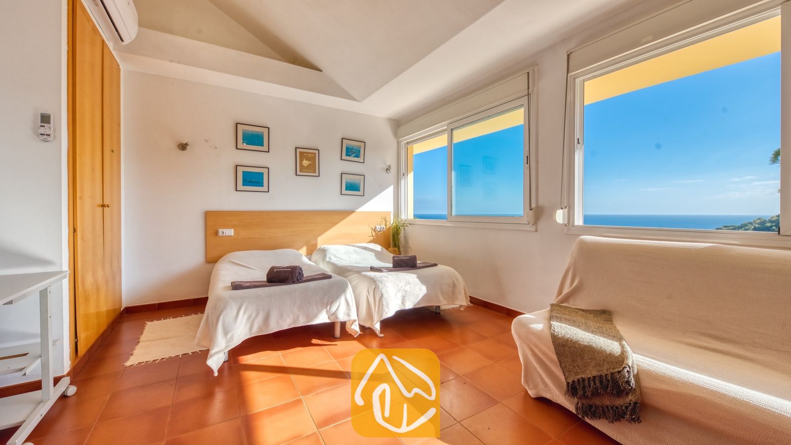 Lichte slaapkamer in Villa Santa Cristina, voorzien van twee eenpersoonsbedden, terracotta tegels en een witte bank. De grote ramen bieden een prachtig uitzicht op de blauwe oceaan onder een heldere hemel.