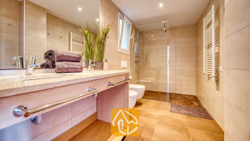 Moderne badkamer met dubbele wastafel, inloopdouche met glazen wand en beige betegelde muren. Opgerolde handdoeken en decoratieve planten sieren het stenen blad.