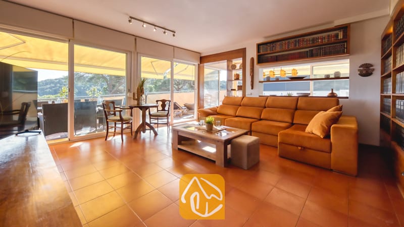 De woonkamer van Villa Santa Cristina is voorzien van een grote oranje hoekbank, terracotta plavuizen en ingebouwde houten boekenplanken naast de glazen schuifdeuren.