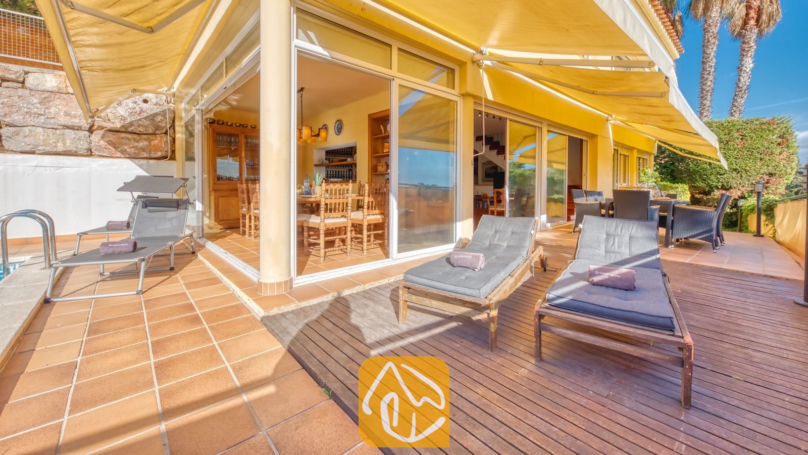 Terras van Villa Santa Cristina, voorzien van houten ligbedden met grijze kussens, grote gele zonneschermen en glazen deuren die toegang bieden tot een eethoek binnen.