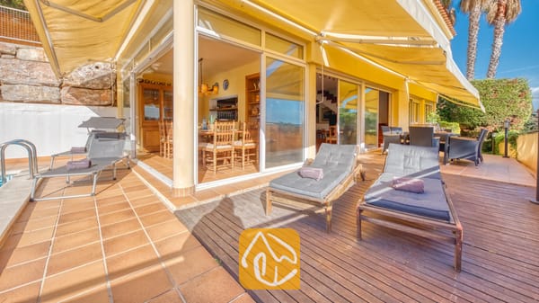 Terras van Villa Santa Cristina, voorzien van houten ligbedden met grijze kussens, grote gele zonneschermen en glazen deuren die toegang bieden tot een eethoek binnen.