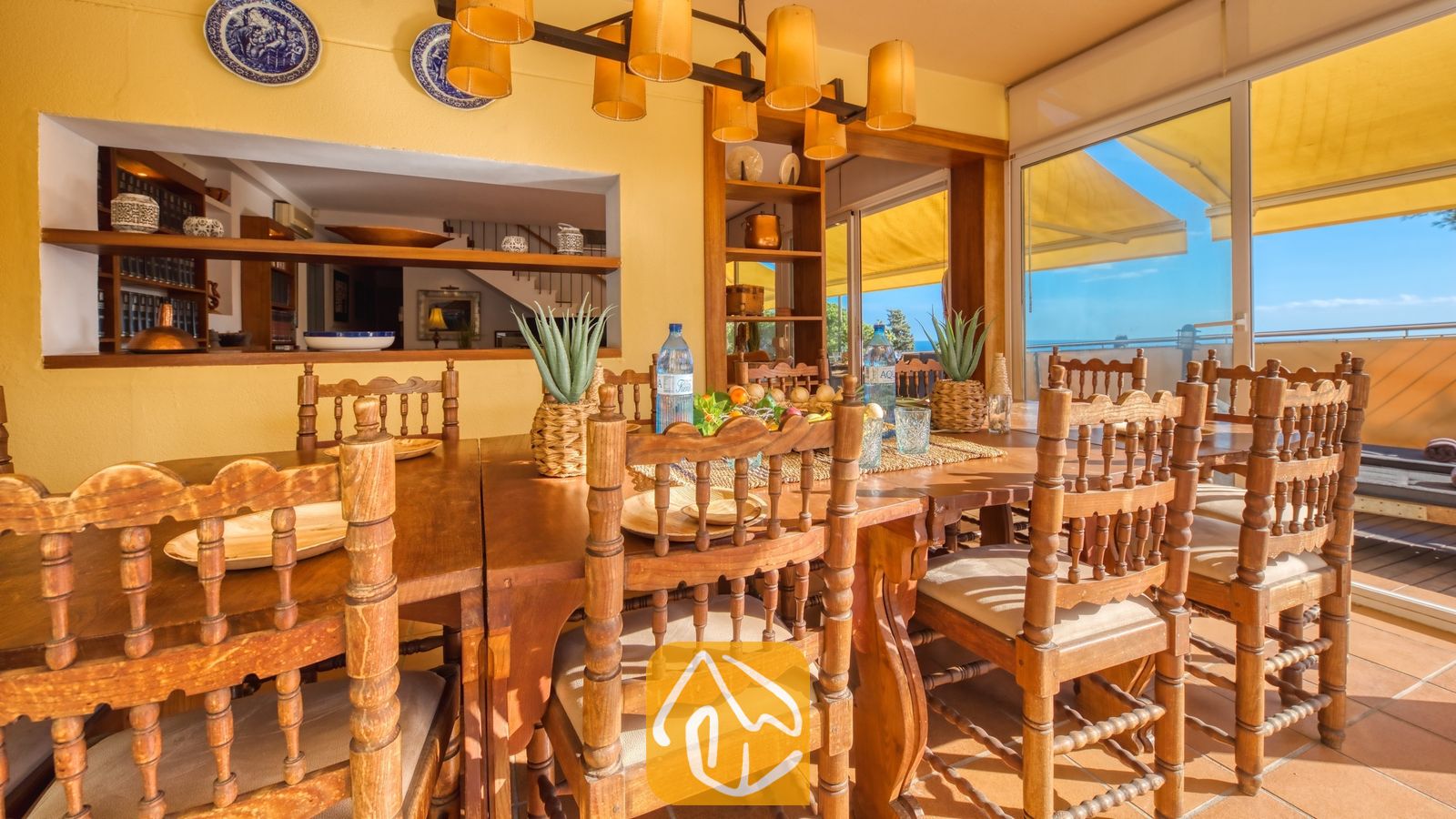 Grote houten eettafel met bewerkte stoelen en crèmekleurige kussens in Villa Santa Cristina. Gele muren, decoratieve blauwe borden en schuifdeuren die toegang bieden tot een zonovergoten terras.
