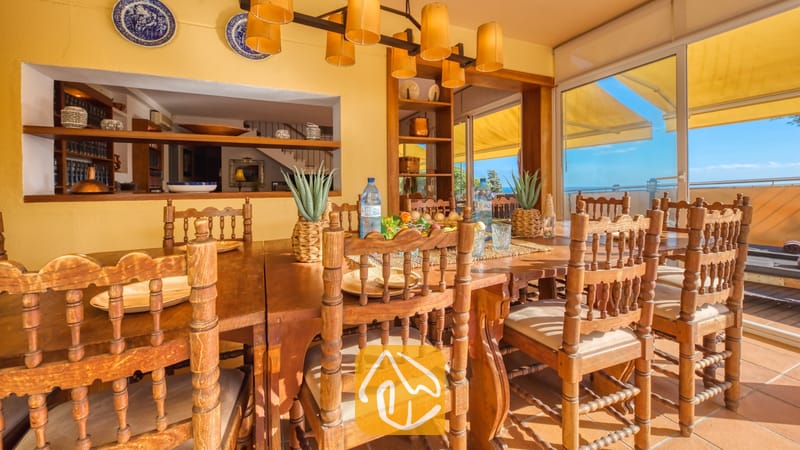 Grote houten eettafel met bewerkte stoelen en crèmekleurige kussens in Villa Santa Cristina. Gele muren, decoratieve blauwe borden en schuifdeuren die toegang bieden tot een zonovergoten terras.