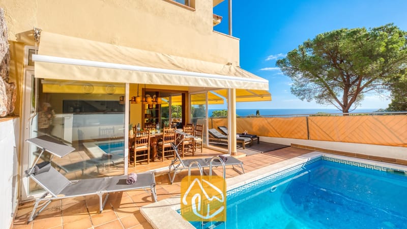 Zwembad en terras bij Villa Santa Cristina met terracotta tegels, grijze ligbedden en een houten eethoek in de schaduw van een geel zonnescherm, met uitzicht op zee.