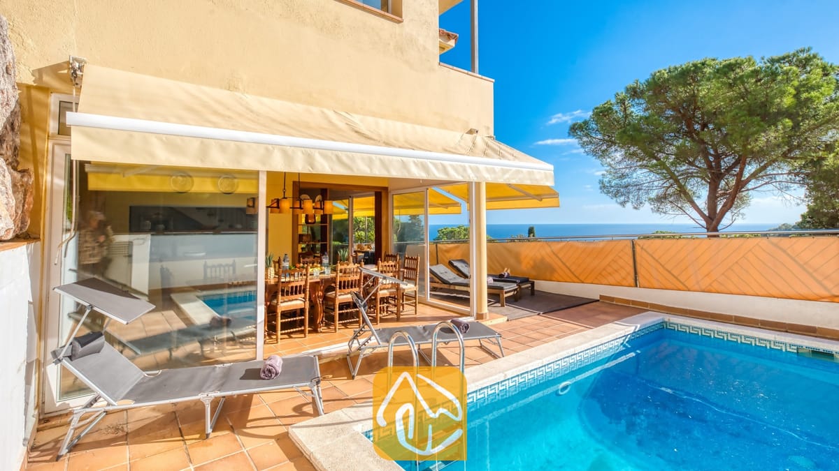 Zwembad en terras bij Villa Santa Cristina met terracotta tegels, grijze ligbedden en een houten eethoek in de schaduw van een geel zonnescherm, met uitzicht op zee.