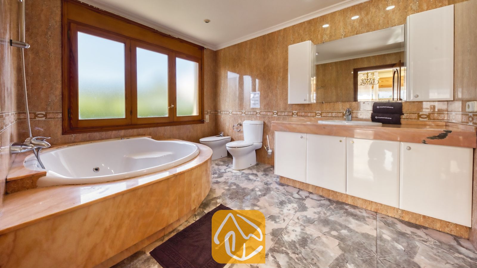 Badkamer voorzien van een hoekbad, witte badkamermeubels, met marmer betegelde muren en een groot raam met een houten kozijn.
