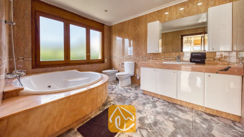 Badkamer voorzien van een hoekbad, witte badkamermeubels, met marmer betegelde muren en een groot raam met een houten kozijn.