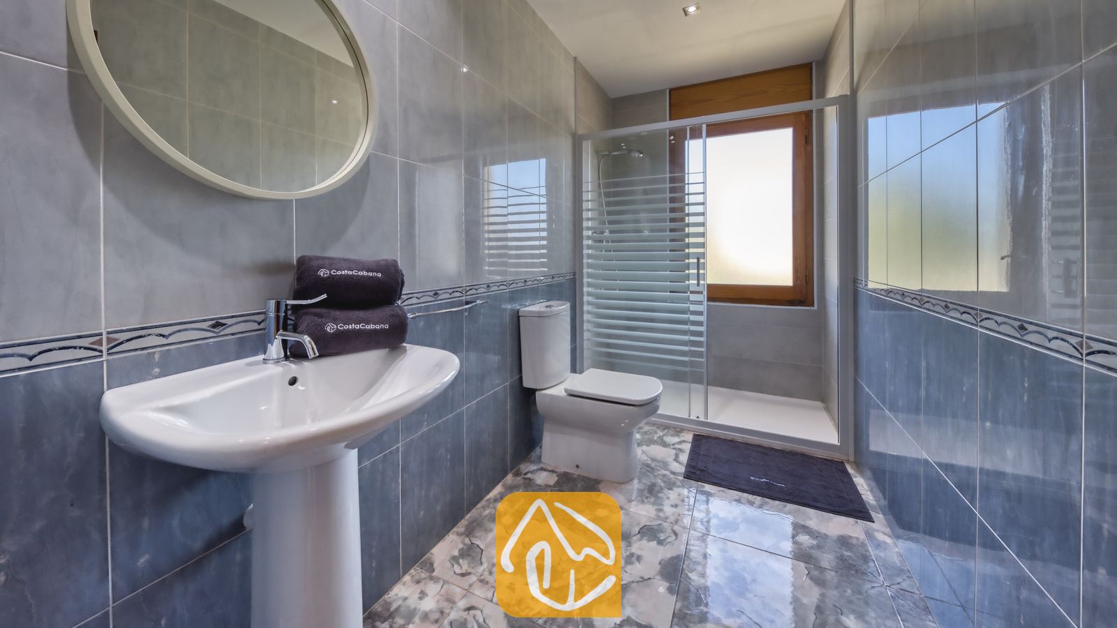 Betegelde badkamer met een wastafel op zuil, een ronde spiegel, een toilet en een glazen doucheruimte met een matglas raam.